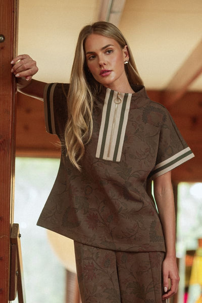 A woman wearing a brown patterned short-sleeve top and matching pants, leaning against a wooden door in a warm indoor setting.