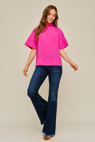 A woman modeling a bright pink short-sleeve mock-neck top with dark blue flared jeans, standing against a plain beige studio background.