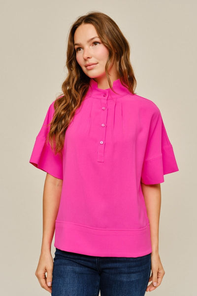 A woman wearing a bright pink short-sleeve blouse with a high mock neck and button placket, paired with dark blue jeans, against a solid light beige background.