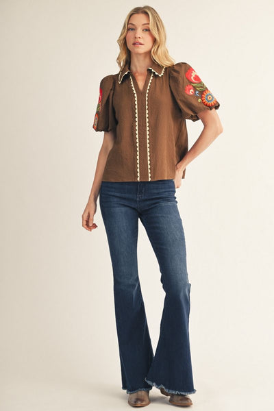 Woman wearing a brown blouse with floral sleeves and blue jeans on a beige background