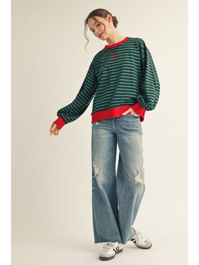 Person wearing a green and red striped sweater with a red shirt underneath, blue jeans, and silver shoes on a plain background.