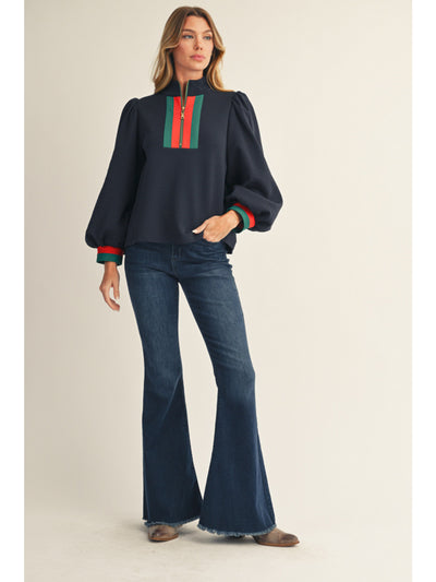 Woman wearing a navy sweater with red and green accents and blue jeans on a beige background