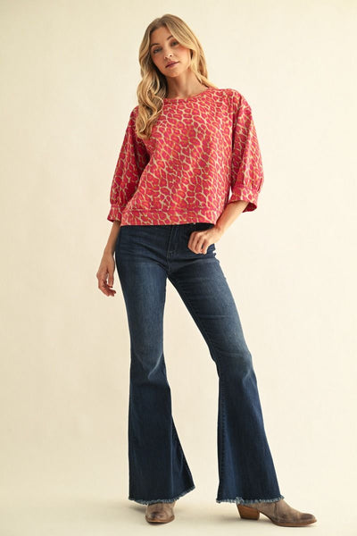 A woman modeling a pink and beige patterned short-sleeve top with dark blue flare jeans and brown boots, standing against a solid beige studio background.