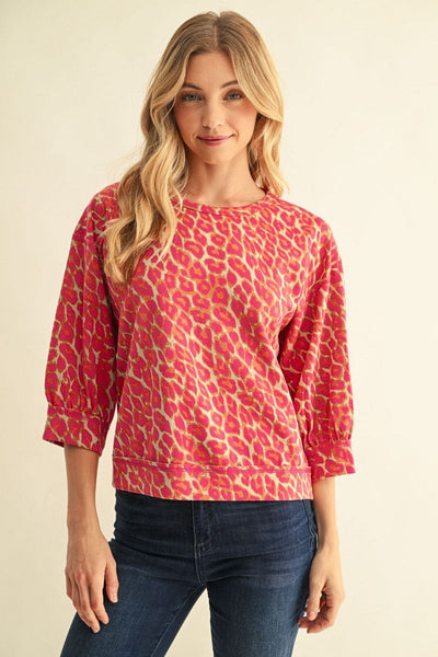 A woman modeling a pink and beige animal print long-sleeve top with three-quarter sleeves, paired with dark blue jeans, against a plain beige background.