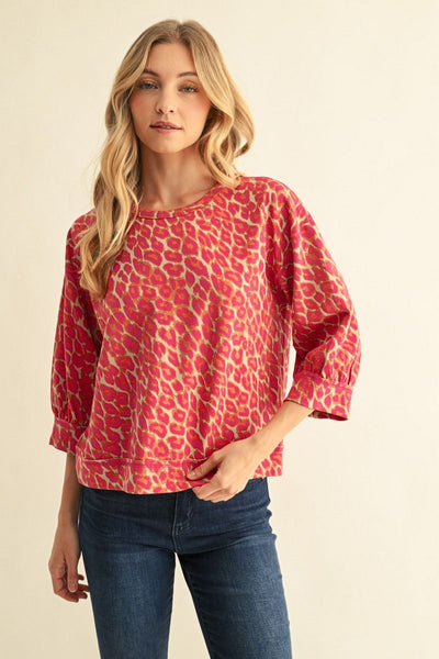 A woman modeling a pink and beige animal print long-sleeve top with three-quarter sleeves, paired with dark blue jeans, against a plain beige background.