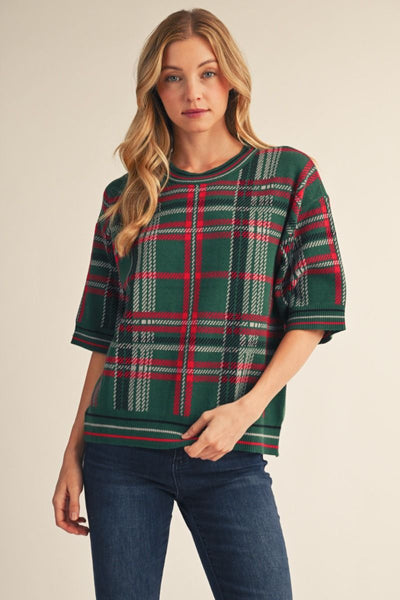 A woman modeling a short-sleeve knit top with a green, red, and white plaid pattern, paired with dark blue jeans, against a solid beige background.