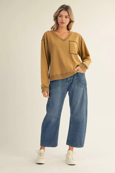 A woman modeling a mustard yellow V-neck long-sleeve sweater with a chest pocket, paired with wide-leg blue jeans and white sneakers, against a plain light beige studio background.