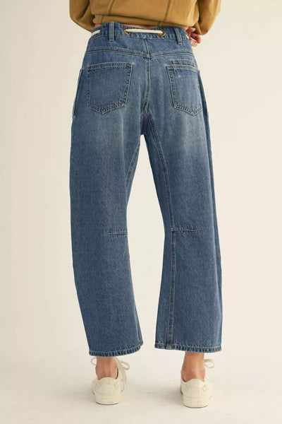 A person shown from the back wearing wide-leg blue denim jeans with a cropped ankle length, paired with a tan top and white sneakers against a plain off-white studio background.