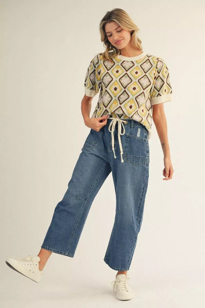 A woman modeling a short-sleeve knit top with a yellow, brown, and white geometric diamond pattern, paired with blue cropped jeans and white sneakers, against a plain light beige studio background.
