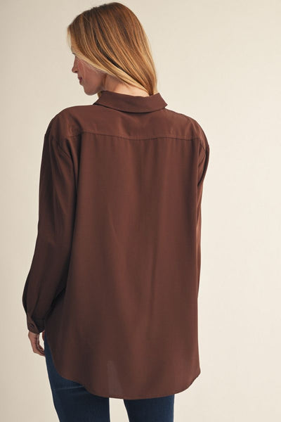 A woman shown from the back wearing an oversized brown button-up shirt and dark blue jeans against a plain light beige studio background.
