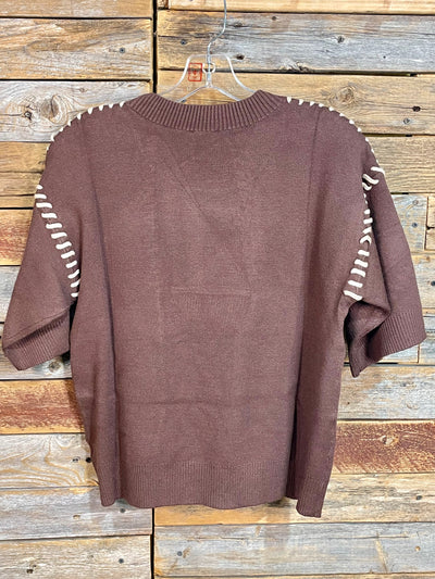 A brown knit short-sleeve sweater with white contrast stitching along the shoulders and sleeves, hanging on a clear plastic hanger against a rustic wooden plank wall.