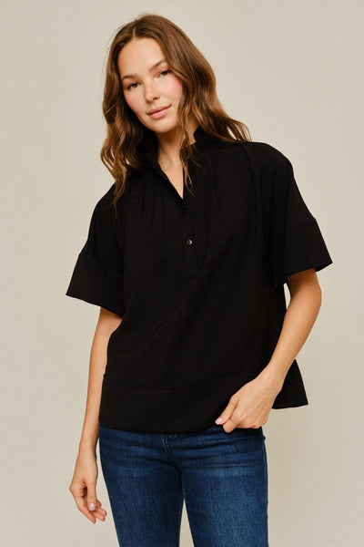 A woman modeling a black short-sleeve button-up shirt with a relaxed, slightly oversized fit and blue jeans, set against a plain light beige studio background.