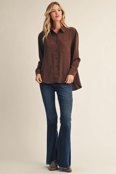 A woman modeling a brown long-sleeve button-up shirt with dark blue flared jeans and brown shoes, standing against a plain beige studio background.