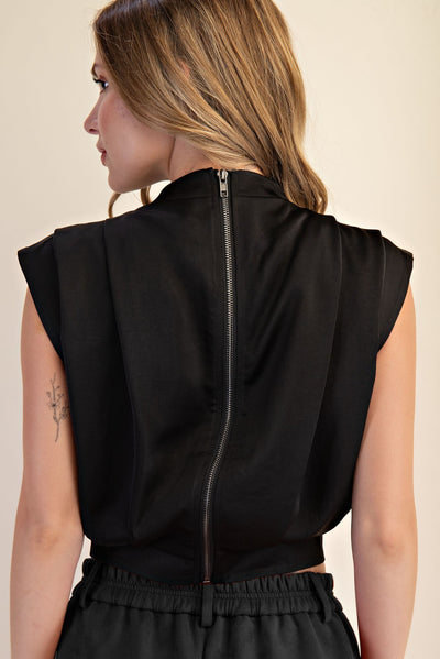 A woman shown from the back wearing a black sleeveless top with a central zipper, against a plain beige studio background.