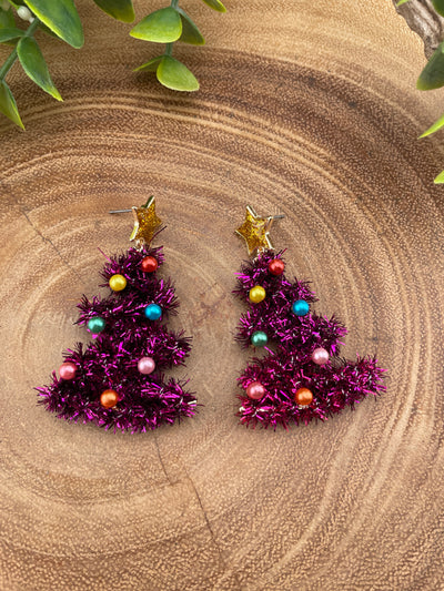 Pink tinsel tree earrings with colorful beads on a textured surface with green leaves.