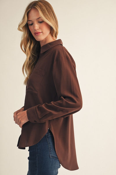 A woman wearing a brown long-sleeve button-up shirt and blue jeans, shown from a side-back angle against a plain light beige studio background.