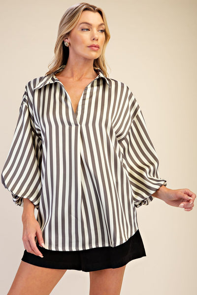 A woman modeling a loose-fitting, oversized button-up blouse with vertical grey and off-white stripes, paired with black shorts, against a solid beige studio background.