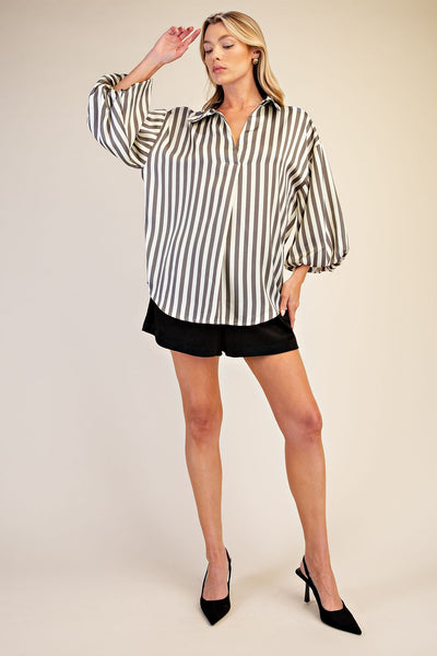 A woman modeling a loose-fitting button-up shirt with verticalgrey and beige stripes, paired with black shorts and black high heels, against a plain beige studio background.