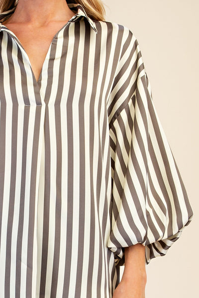 A close-up of a woman wearing a loose-fitting collared blouse with vertical grey and off-white stripes, featuring voluminous elbow-length sleeves.