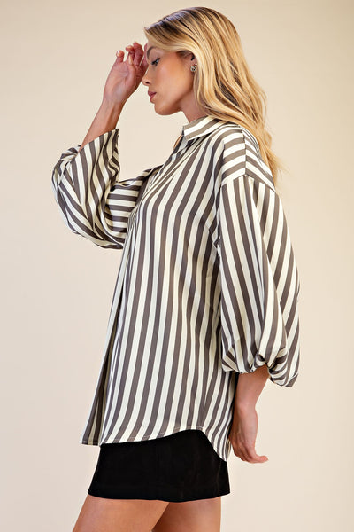 A woman wearing a loose, oversized long-sleeve blouse with vertical grey and cream stripes, paired with a black skirt, posed against a solid beige studio background.