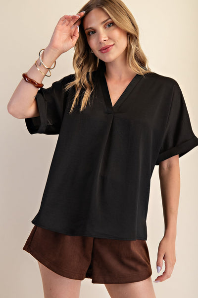 A woman wearing a loose black short-sleeve V-neck blouse with rolled cuffs and brown shorts, posed against a plain beige studio background.
