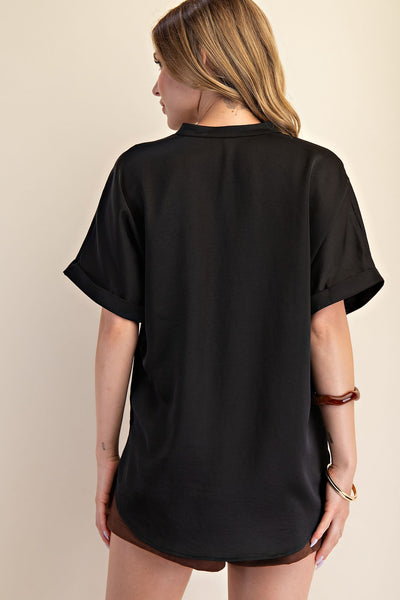 A woman shown from the back wearing an oversized black short-sleeve top with rolled cuffs, photographed against a plain beige studio background.