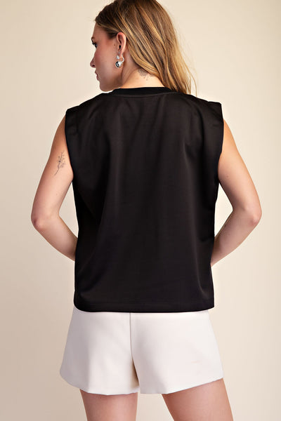 A woman shown from the back wearing a black sleeveless top and white shorts against a plain beige studio background.