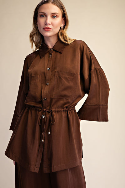 A woman modeling a brown long-sleeve collared shirt dress with a button front, chest pockets, and a drawstring waist, photographed against a plain beige studio background.