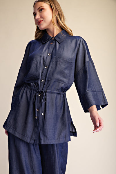 A woman wearing a dark blue denim-like button-up shirt with a tie waist and matching wide-leg pants, photographed against a plain beige studio background.