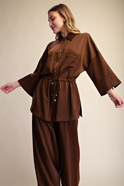 A woman modeling a brown two-piece outfit consisting of a button-front long-sleeve top with a drawstring waist and matching wide-leg pants, photographed against a plain beige studio background.