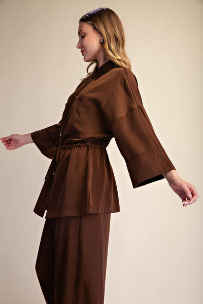 A woman wearing a matching brown two-piece outfit, featuring a cropped button-front jacket with wide three-quarter sleeves and matching wide-leg trousers, posed against a solid beige studio background.