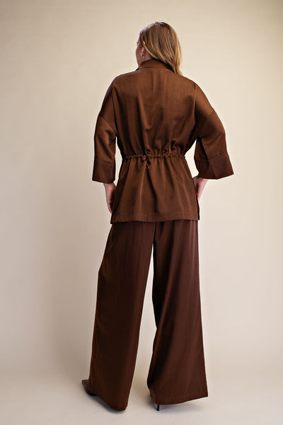 A woman viewed from the back wearing a matching brown two-piece outfit consisting of a cropped, 3/4 sleeve belted jacket and wide-leg trousers, set against a plain beige studio background.