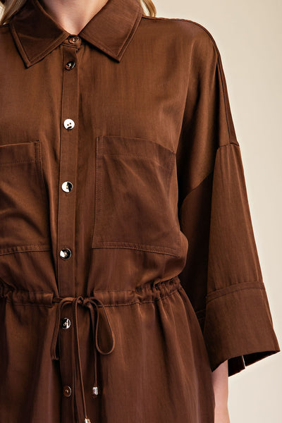 A close-up of a model wearing a brown collared shirt dress with silver buttons, chest pockets, and a drawstring waist against a light beige background.