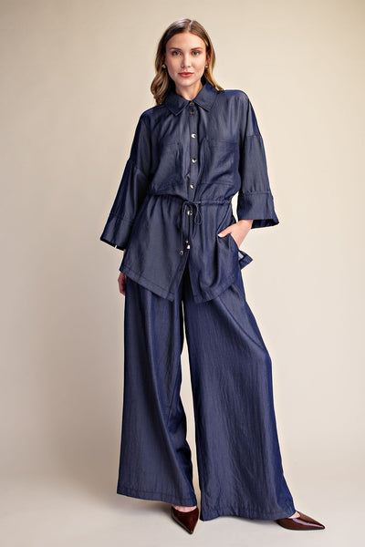 A woman modeling a blue two-piece set consisting of a 3/4 sleeve button up shirt and matching wide-leg pants, photographed against a solid beige studio background.