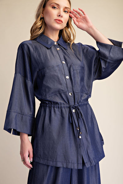 A woman wearing a blue button-front shirt dress with wide three-quarter sleeves, a pointed collar, and a drawstring waist, photographed against a plain beige studio background.