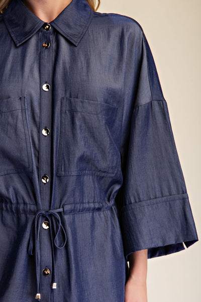 A close-up of a blue shirt with a collar, button-down front, chest pockets, and a drawstring waist, shown on a model against a light beige background.