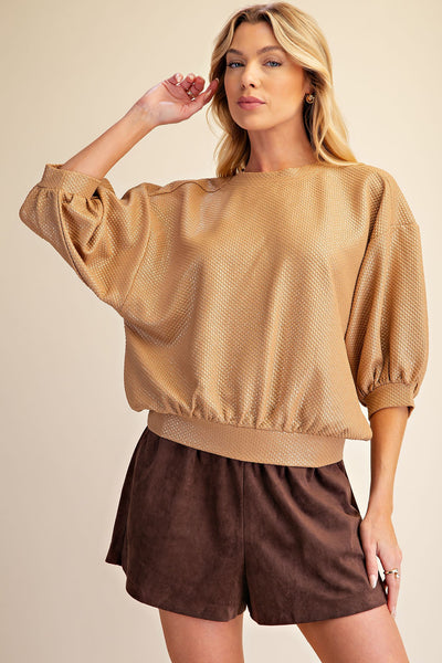 A woman modeling a textured gold long-sleeve top with voluminous puff sleeves and dark brown shorts against a solid beige background.