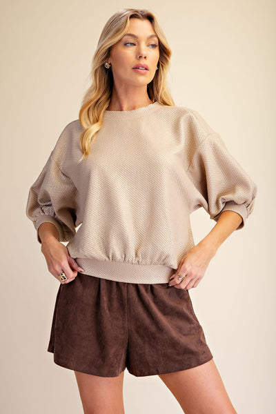 A woman modeling a beige textured bubble-sleeve sweatshirt and dark brown shorts against a solid beige studio background.