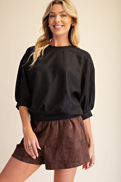 A smiling blonde woman wearing a black textured long-sleeve top with puff sleeves and brown shorts, standing against a plain beige studio background.