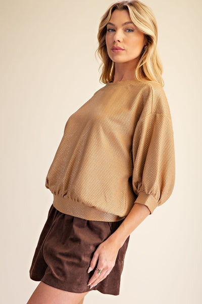 A woman modeling a textured, waffle-knit long-sleeve top in camel beige with voluminous balloon sleeves and dark brown shorts, set against a solid warm beige background.