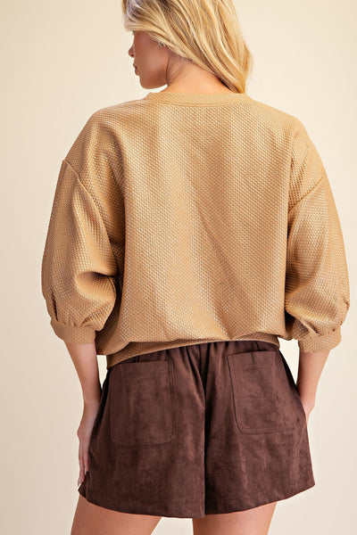 A woman shown from the back wearing a textured gold long-sleeve top with puffed sleeves and dark brown shorts against a plain beige background.