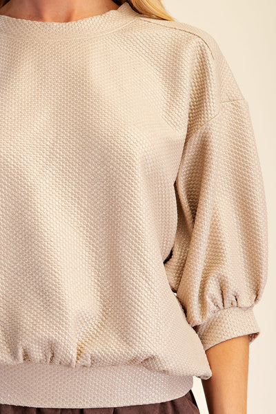 Close-up of a person wearing a textured cream-colored long-sleeve top with a waffle or honeycomb fabric pattern, featuring a crew neck and gathered cuffs.