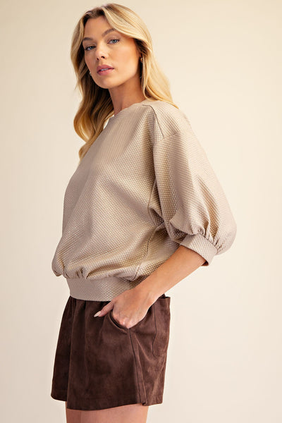A woman wearing a textured beige bubble-sleeve top and a brown skirt, posing against a solid light beige background.