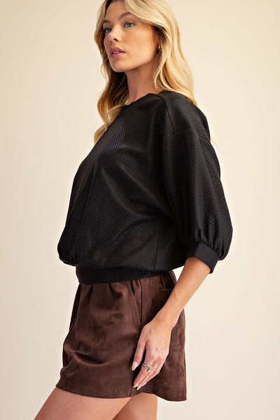 A woman modeling a black textured puff-sleeve top and brown shorts against a warm beige studio background.