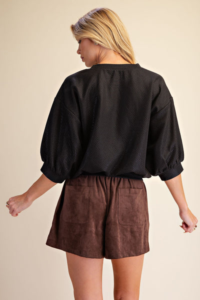 A woman shown from the back wearing a black textured long-sleeve top with puff sleeves and brown shorts, set against a solid beige studio background.