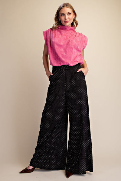 A woman modeling a bright pink sleeveless mock-neck top paired with black wide-leg pants featuring rhinestones, set against a plain beige studio background.