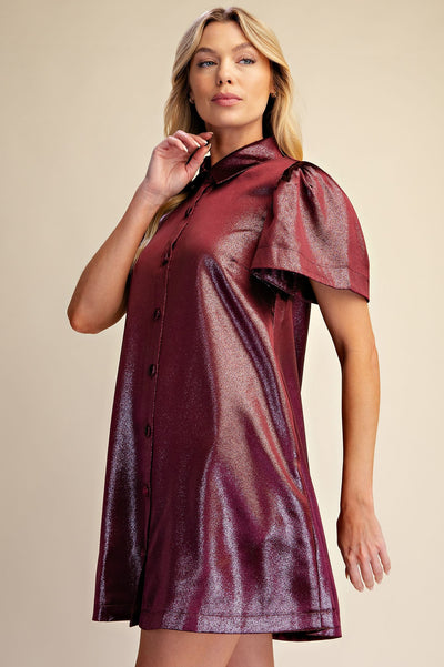 A woman modeling a metallic burgundy mini dress with a high collar, front buttons, and dramatic ruffled sleeves against a plain beige studio background.