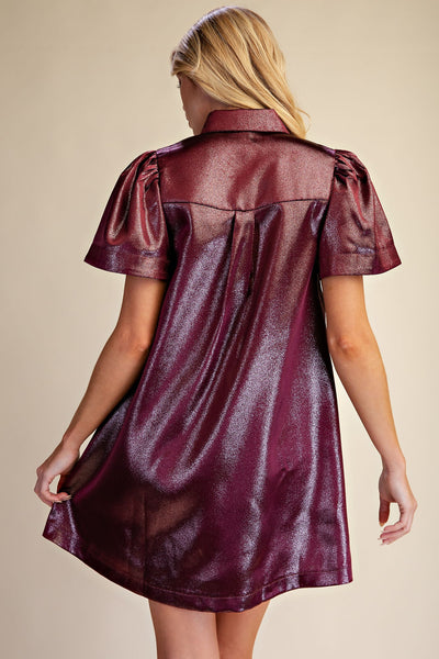 A woman shown from the back wearing a short-sleeve metallic burgundy shirt dress with a glossy finish, set against a plain beige background.