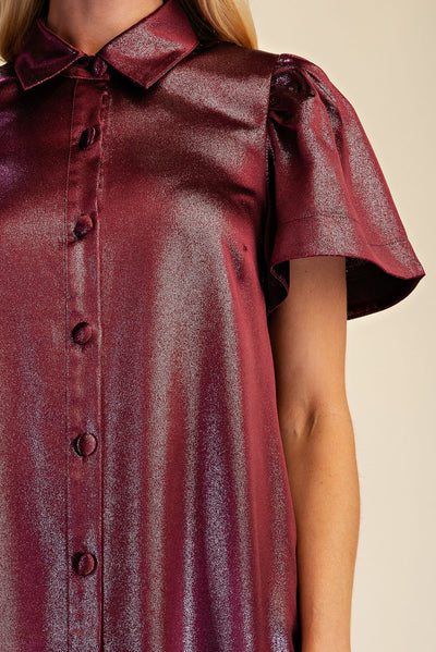 A close-up of a model wearing a short-sleeve collared shirt dress in a metallic burgundy fabric with a glossy finish, set against a plain beige background.