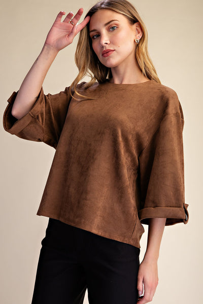 A woman wearing a brown suede-like cropped top with three-quarter sleeves and black pants, posed against a plain beige studio background.